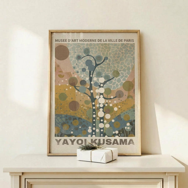 May include: Framed poster in natural wood frame leaning against white wall on light wooden surface. Features abstract tree design with circular dots in muted greens, blues, and earth tones. Text reads "Musee d'Art Moderne de la Ville de Paris" and "Yayoi Kusama" at bottom. Small white wrapped gift with green sprig sits beside frame. Minimalist room setting with neutral colors.