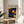 May include: Framed poster with blue background featuring yellow and orange circular dots in Kusama's signature polka dot style, displayed on light wood round side table next to beige upholstered chair. Natural sunlight creates soft shadows on white wall. Green plant leaves visible in corner. Artwork shows Japanese text and "KYOTO" with abstract spotted pattern resembling celestial bodies against deep blue field.