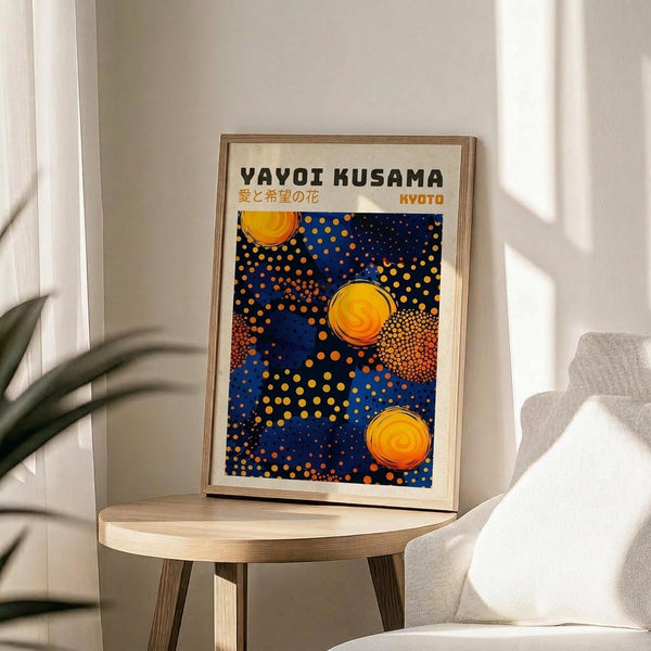 May include: Framed poster with blue background featuring yellow and orange circular dots in Kusama's signature polka dot style, displayed on light wood round side table next to beige upholstered chair. Natural sunlight creates soft shadows on white wall. Green plant leaves visible in corner. Artwork shows Japanese text and "KYOTO" with abstract spotted pattern resembling celestial bodies against deep blue field.