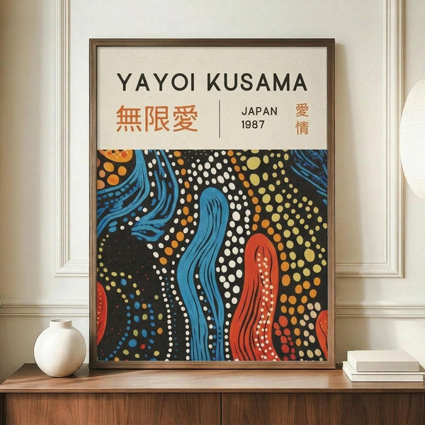 May include: Framed art print featuring Yayoi Kusama's distinctive polka dot style with flowing blue and red organic shapes against black background filled with white, orange and yellow dots. The poster displays "YAYOI KUSAMA" at top with Japanese characters and "JAPAN 1987" text. Wooden frame sits on light wood console table beside white ceramic vase and stacked books against cream wall with white molding.
