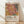 May include: Framed poster print leaning against white wall on light wooden floor. Abstract botanical artwork features coral pink monstera leaf with yellow veining, surrounded by tropical foliage in earth tones including spotted patterns and palm fronds. Text shows "YAYOI KUSAMA" with Japanese characters and "MUSÉE D'ART MODERNE DE LA VILLE DE PARIS" on cream background. Natural wood frame with soft window light casting shadows across white textured wall.