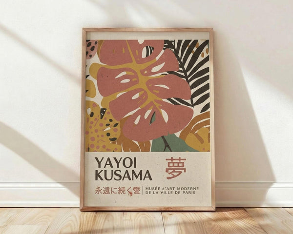 May include: Framed poster print leaning against white wall on light wooden floor. Abstract botanical artwork features coral pink monstera leaf with yellow veining, surrounded by tropical foliage in earth tones including spotted patterns and palm fronds. Text shows "YAYOI KUSAMA" with Japanese characters and "MUSÉE D'ART MODERNE DE LA VILLE DE PARIS" on cream background. Natural wood frame with soft window light casting shadows across white textured wall.