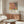 May include: Modern living room mockup showing framed art print on white wall above wooden sideboard. Poster features abstract botanical design with coral pink monstera leaf, yellow and black tropical foliage, and Japanese text. Room includes white curved chair, glass oval coffee table, brass floor lamp, and neutral decor with natural lighting.