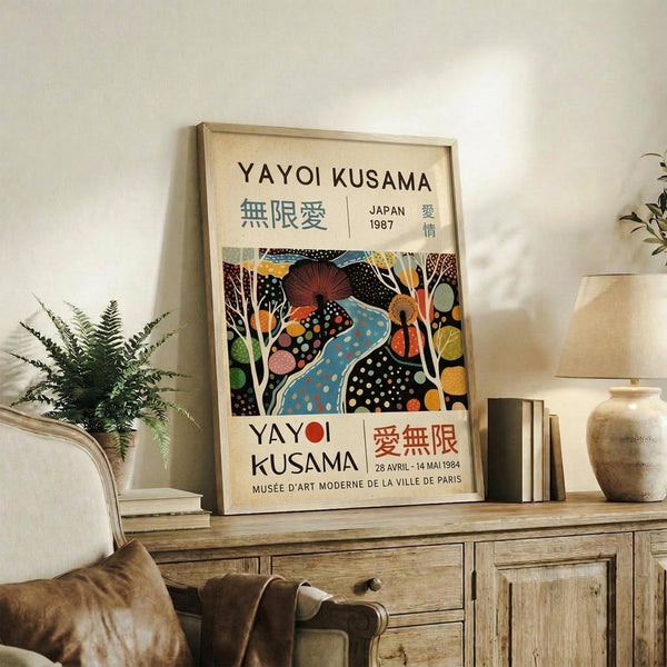 May include: Framed vintage-style exhibition poster featuring Yayoi Kusama artwork with colorful polka-dotted abstract shapes, trees, and organic forms in blues, reds, yellows, and greens. Text in Japanese and French indicates museum exhibition dates. Poster displayed on rustic wooden sideboard alongside fern plant, ceramic vase, books, and cream upholstered furniture in neutral-toned living room setting.