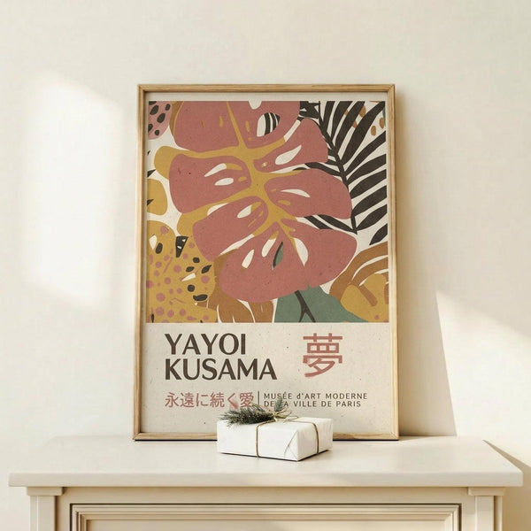 May include: Framed art print featuring abstract botanical design with coral pink monstera leaf, yellow and black patterned foliage, and striped palm fronds on cream background. Text shows "YAYOI KUSAMA" with Japanese characters and museum attribution. Natural wood frame displayed on light wooden surface with small wrapped gift box. Warm neutral tones throughout minimalist setting.