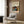 May include: Framed Yayoi Kusama exhibition poster displayed on wood-paneled wall above beige upholstered armchair with geometric pillow. Poster features colorful abstract artwork with polka dots and organic shapes in blues, oranges, and earth tones against cream background. Modern living room setting with brass floor lamp, dark round side table with open book, and natural lighting from window with sheer curtains.