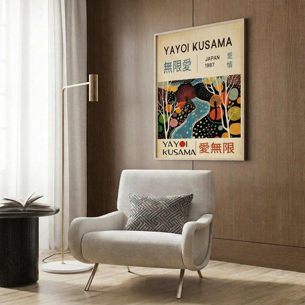 May include: Framed Yayoi Kusama exhibition poster displayed on wood-paneled wall above beige upholstered armchair with geometric pillow. Poster features colorful abstract artwork with polka dots and organic shapes in blues, oranges, and earth tones against cream background. Modern living room setting with brass floor lamp, dark round side table with open book, and natural lighting from window with sheer curtains.