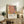 May include: Framed art print with abstract botanical design featuring coral pink monstera leaf, yellow and green tropical foliage, and black zebra-striped palm frond against cream background. Text reads "YAYOI KUSAMA" with Japanese characters and "MUSÉE D'ART MODERNE DE LA VILLE DE PARIS" below. Print displayed on rustic wooden sideboard alongside potted fern, ceramic vase, and books in neutral-toned living room setting.