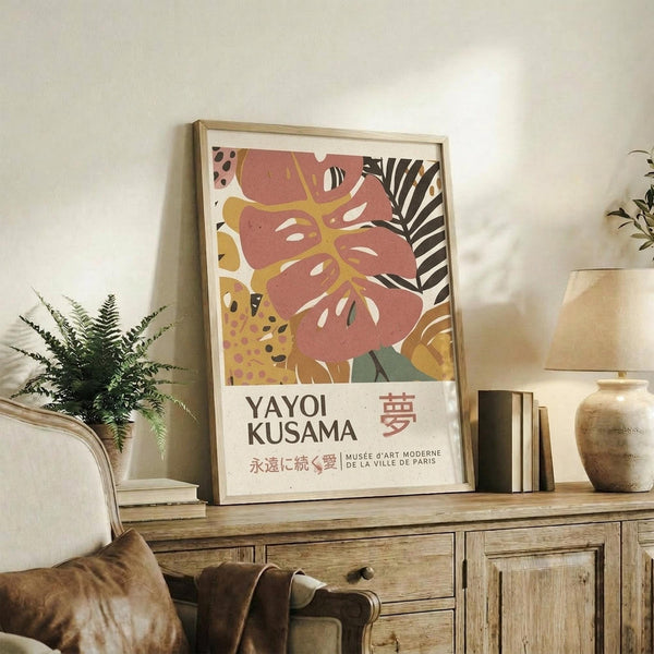 May include: Framed art print with abstract botanical design featuring coral pink monstera leaf, yellow and green tropical foliage, and black zebra-striped palm frond against cream background. Text reads "YAYOI KUSAMA" with Japanese characters and "MUSÉE D'ART MODERNE DE LA VILLE DE PARIS" below. Print displayed on rustic wooden sideboard alongside potted fern, ceramic vase, and books in neutral-toned living room setting.