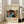 May include: Framed vintage-style exhibition poster leaning against white wall next to beige sofa in bright room with large windows. Poster features colorful abstract art with polka dots, flowing blue river, stylized trees, and organic shapes in vibrant yellows, oranges, and greens on black background. Text includes artist name, Japanese characters, museum details, and exhibition dates. Light wood frame on hardwood floor with area rug.