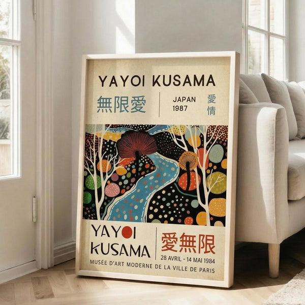 May include: Framed vintage-style exhibition poster leaning against white wall next to beige sofa in bright room with large windows. Poster features colorful abstract art with polka dots, flowing blue river, stylized trees, and organic shapes in vibrant yellows, oranges, and greens on black background. Text includes artist name, Japanese characters, museum details, and exhibition dates. Light wood frame on hardwood floor with area rug.