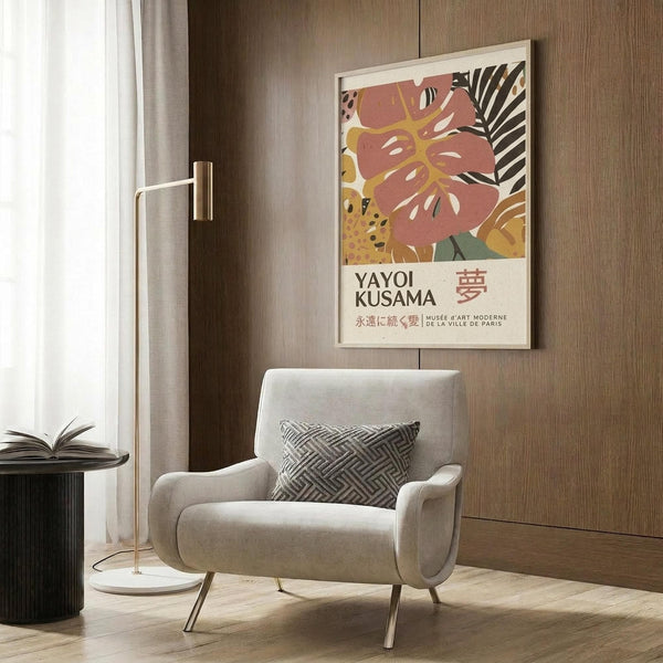May include: Modern interior with beige armchair featuring geometric patterned pillow, brass floor lamp, dark round side table with open book. Framed poster on wood-paneled wall shows abstract tropical leaves in coral pink, yellow, and black with "YAYOI KUSAMA" text and Japanese characters below. Neutral curtains visible at left, hardwood flooring throughout the contemporary living space.