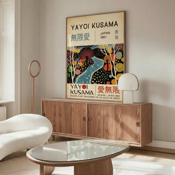 May include: Colorful Yayoi Kusama exhibition poster mounted above wooden credenza in modern living room. Poster features abstract organic shapes in blues, oranges, and earth tones with polka dot patterns and Japanese text. Room has white curved chair, brass floor lamp, oval glass coffee table, and neutral walls with natural lighting.