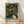 May include: Framed poster print leaning against white wall on light wood floor. Features abstract art with swirling dark green background filled with colorful circles and dots in yellow, orange, red, pink, and blue tones. White polka-dotted organic shape in lower portion. Text reads "YAYOI KUSAMA" with Japanese characters and "TOKYO" in red. Natural wood frame, soft window light casting shadows on textured white wall.