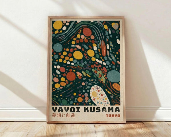 May include: Framed poster print leaning against white wall on light wood floor. Features abstract art with swirling dark green background filled with colorful circles and dots in yellow, orange, red, pink, and blue tones. White polka-dotted organic shape in lower portion. Text reads "YAYOI KUSAMA" with Japanese characters and "TOKYO" in red. Natural wood frame, soft window light casting shadows on textured white wall.