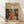 May include: Framed art print leaning against white wall on light wood floor. Wooden frame contains beige matted poster featuring colorful abstract pattern with polka dots, organic shapes, and floral motifs in orange, blue, red, and yellow. Black text at top reads "YAYOI KUSAMA" with Japanese characters and "JAPAN 1987". Soft natural lighting creates diagonal shadows across white fabric background.