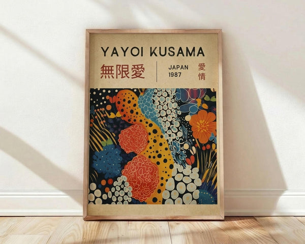 May include: Framed art print leaning against white wall on light wood floor. Wooden frame contains beige matted poster featuring colorful abstract pattern with polka dots, organic shapes, and floral motifs in orange, blue, red, and yellow. Black text at top reads "YAYOI KUSAMA" with Japanese characters and "JAPAN 1987". Soft natural lighting creates diagonal shadows across white fabric background.