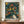 May include: Framed art print displayed on wooden sideboard featuring abstract design with dark teal background filled with colorful circles, dots, and flowing patterns in yellow, orange, red, and blue. Text reads "YAYOI KUSAMA" with Japanese characters and "TOKYO" below. White ceramic vase and stacked books sit alongside the medium-sized framed poster against cream colored walls with white molding.