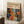 May include: Framed art poster with cream background and colorful abstract pattern featuring polka dots, floral motifs, and organic shapes in orange, blue, red, and white. Text reads "YAYOI KUSAMA" with Japanese characters and "JAPAN 1987". Light wood frame leaning against white wall corner, white coffee cup on wooden block nearby, beige textured rug beneath.