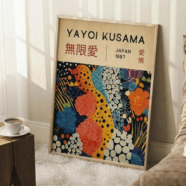May include: Framed art poster with cream background and colorful abstract pattern featuring polka dots, floral motifs, and organic shapes in orange, blue, red, and white. Text reads "YAYOI KUSAMA" with Japanese characters and "JAPAN 1987". Light wood frame leaning against white wall corner, white coffee cup on wooden block nearby, beige textured rug beneath.