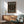 May include: Framed poster with black background and colorful polka dots in muted tones of coral, sage green, cream, and peach, displayed above a wooden sideboard with white ceramic lamp. Mid-century modern teal upholstered chair with wood arms sits beside small round coffee table. Warm natural lighting creates shadows on white wall in minimalist living room setting.