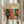 May include: Person holding framed art print featuring colorful abstract pattern with polka dots, organic shapes, and floral motifs in orange, blue, yellow, and red against dark background. Print has cream mat with black text including Japanese characters. Light wood frame, neutral beige clothing, dried pampas grass visible, soft natural lighting near window with white curtains.