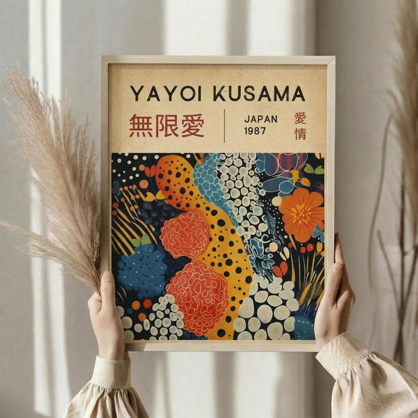 May include: Person holding framed art print featuring colorful abstract pattern with polka dots, organic shapes, and floral motifs in orange, blue, yellow, and red against dark background. Print has cream mat with black text including Japanese characters. Light wood frame, neutral beige clothing, dried pampas grass visible, soft natural lighting near window with white curtains.