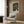 May include: Colorful abstract art poster with swirling dark green background filled with circles in yellow, orange, red, and teal, displayed in light wood frame on textured wood wall panel. Modern beige armchair with geometric patterned pillow sits below artwork, alongside brass floor lamp and dark round side table with open book. Neutral curtains visible on left side.
