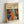 May include: Framed poster print leaning against white wall corner featuring colorful abstract pattern with organic shapes, polka dots, and cellular forms in orange, blue, red, and yellow against dark background. Light wood frame surrounds cream-colored mat with "YAYOI KUSAMA" text and Japanese characters at top, plus "JAPAN 1987" marking. Artwork displays vibrant, psychedelic style with flowing, interconnected forms and varied textures.
