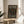 May include: A framed poster with black background featuring colorful polka dots in muted tones of peach, sage green, cream, and coral. White text reads "YAYOI KUSAMA INFINITY DOTS FREVER" at top and "INFINITY DOTS FOREVER MUSEUM OF CONTEMPORARY ART 22 - APRIL 1986" at bottom. The thin wooden frame sits on a round wooden side table next to a beige upholstered chair, with plant leaves and natural window light visible in a minimalist room setting.