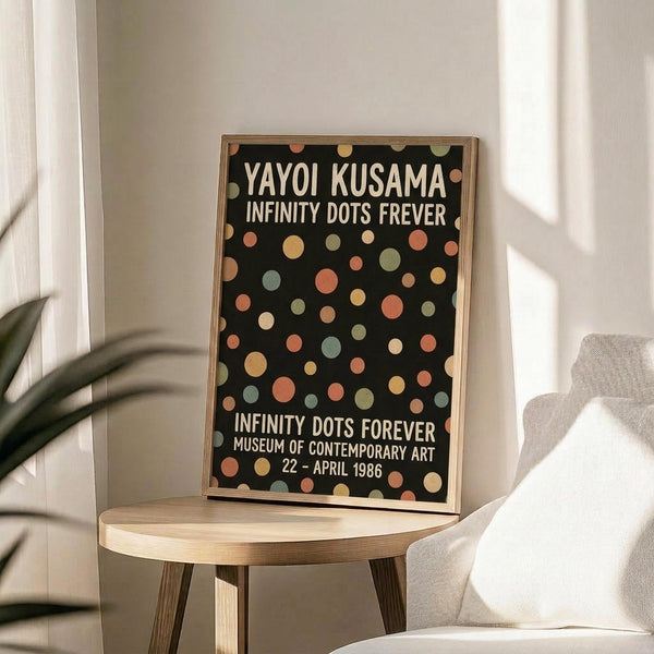 May include: A framed poster with black background featuring colorful polka dots in muted tones of peach, sage green, cream, and coral. White text reads "YAYOI KUSAMA INFINITY DOTS FREVER" at top and "INFINITY DOTS FOREVER MUSEUM OF CONTEMPORARY ART 22 - APRIL 1986" at bottom. The thin wooden frame sits on a round wooden side table next to a beige upholstered chair, with plant leaves and natural window light visible in a minimalist room setting.