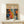 May include: Framed art print displayed on light wood shelf against cream wall. Poster features colorful abstract pattern with polka dots, cellular shapes, and organic forms in orange, blue, red, and white. Text reads "YAYOI KUSAMA" with Japanese characters and "JAPAN 1987". Small white gift box with evergreen sprig sits beside the natural wood frame.