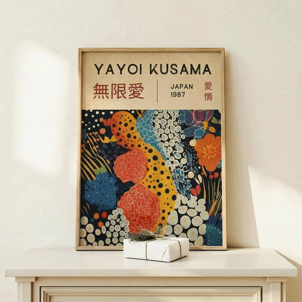 May include: Framed art print displayed on light wood shelf against cream wall. Poster features colorful abstract pattern with polka dots, cellular shapes, and organic forms in orange, blue, red, and white. Text reads "YAYOI KUSAMA" with Japanese characters and "JAPAN 1987". Small white gift box with evergreen sprig sits beside the natural wood frame.
