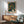 May include: Framed poster with abstract organic pattern in dark green featuring colorful dots, circles and flowing lines in yellow, orange, white and teal, displayed above mid-century modern wood credenza with white ceramic lamp, next to mint green upholstered armchair with wood frame in bright sunlit room with white walls.