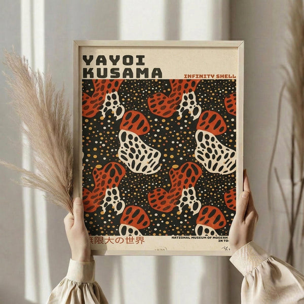 May include: Person holding framed art print featuring repeating pattern of polka-dotted mushrooms in red, white, and black colors on dark background with yellow dots. Light wooden frame, cream matting. Person wears beige clothing, holds dried pampas grass stems. Soft natural lighting against neutral curtained background.