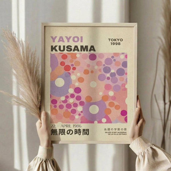 May include: Person holding framed art poster featuring colorful polka dot pattern in pink, purple, orange and cream tones. Text reads "Yayoi Kusama Tokyo 1998" and Japanese characters. Light wooden frame against neutral curtained background with dried pampas grass visible on left side. Person wearing beige ruffled sleeve top.
