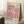 May include: Framed exhibition poster leaning against white wall corner featuring colorful overlapping circles in pink, purple, orange and cream tones. Text reads "YAYOI KUSAMA" and "TOKYO 1998" with Japanese characters below. Natural wood frame, positioned on white textured rug next to wooden block with coffee cup. Soft natural lighting creates shadows on wall.