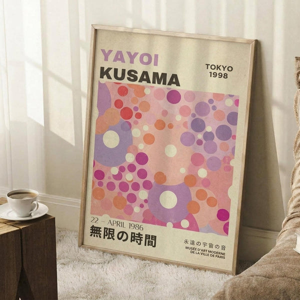 May include: Framed exhibition poster leaning against white wall corner featuring colorful overlapping circles in pink, purple, orange and cream tones. Text reads "YAYOI KUSAMA" and "TOKYO 1998" with Japanese characters below. Natural wood frame, positioned on white textured rug next to wooden block with coffee cup. Soft natural lighting creates shadows on wall.