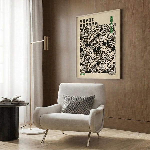 May include: Framed Yayoi Kusama exhibition poster with black polka dot pattern on cream background, displayed above light gray upholstered armchair with geometric patterned pillow. Modern minimalist room setting features brass floor lamp, dark round side table with open book, warm wood paneled walls, and white curtains. Natural lighting creates soft shadows on light wooden flooring.