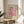 May include: Framed art print featuring colorful polka dot pattern in pink, purple, orange and cream tones, displayed on round wooden side table next to beige upholstered chair. Print shows "YAYOI KUSAMA" text at top with Japanese characters below and "TOKYO 1998" in corner. Natural sunlight streams across white wall creating soft shadows from nearby plant leaves.