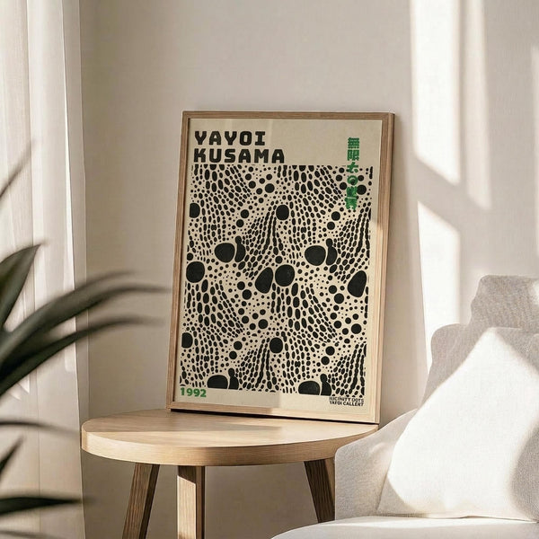 May include: Framed poster with black polka dot pattern on cream background, displaying "YAYOI KUSAMA" text and "1992" in green, propped on light wood round side table next to beige upholstered chair with decorative pillow, green plant leaves visible in corner, natural sunlight casting shadows on white wall
