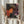 May include: Person in cream sweater holding framed poster featuring colorful polka dot pattern with flower-like shapes in orange, red, pink against dark background. Japanese text at bottom. Dried pampas grass visible to left. Light wooden frame, neutral wall background.