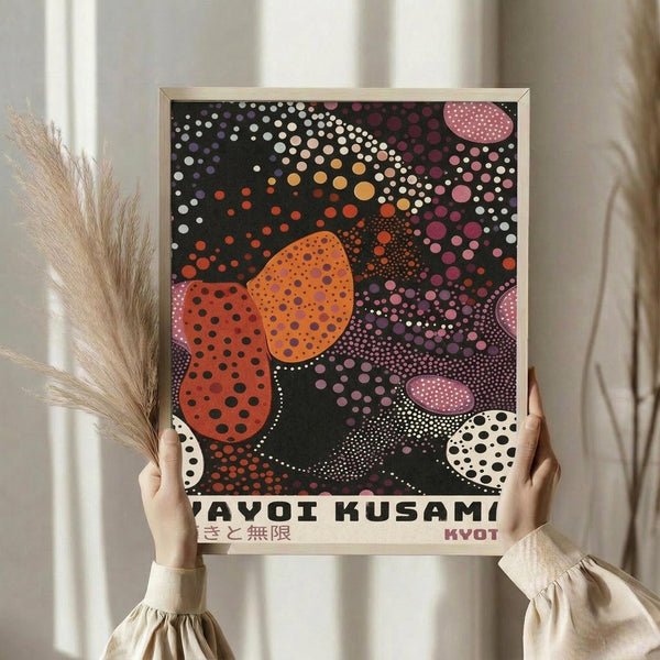 May include: Person in cream sweater holding framed poster featuring colorful polka dot pattern with flower-like shapes in orange, red, pink against dark background. Japanese text at bottom. Dried pampas grass visible to left. Light wooden frame, neutral wall background.