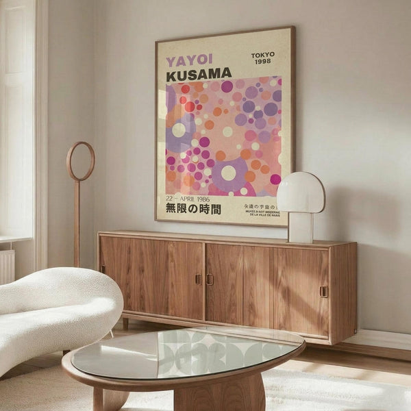 May include: Colorful polka dot art poster in thin frame hanging above wooden credenza in modern living room. Artwork features purple, pink, orange and white circles on cream background with "YAYOI KUSAMA" and "TOKYO 1998" text. Room has white curved chair, glass oval coffee table, floor lamp, and neutral walls with natural lighting.