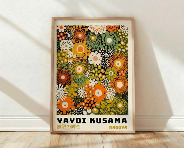 May include: Framed poster with colorful abstract floral pattern featuring overlapping circular and daisy-like shapes in orange, green, yellow, white, and black dots. The artwork displays "YAYOI KUSAMA" and Japanese text with "NAGOYA" below. Light wood frame sits on wooden floor against white textured wall with soft natural lighting creating gentle shadows.