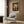 May include: Colorful framed art print featuring a silhouetted tree with vibrant rainbow foliage and scattered dots against a cosmic background, displayed above a cream upholstered armchair with geometric throw pillow in a modern living room with wood paneling, brass floor lamp, and side table with open book.