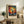 May include: Framed art print featuring a vibrant tree with colorful dotted foliage in reds, yellows, blues and greens against a cosmic background, displayed on a rustic wooden sideboard next to a fern plant, ceramic vase, and books in a neutral-toned living room setting with cream chair visible.