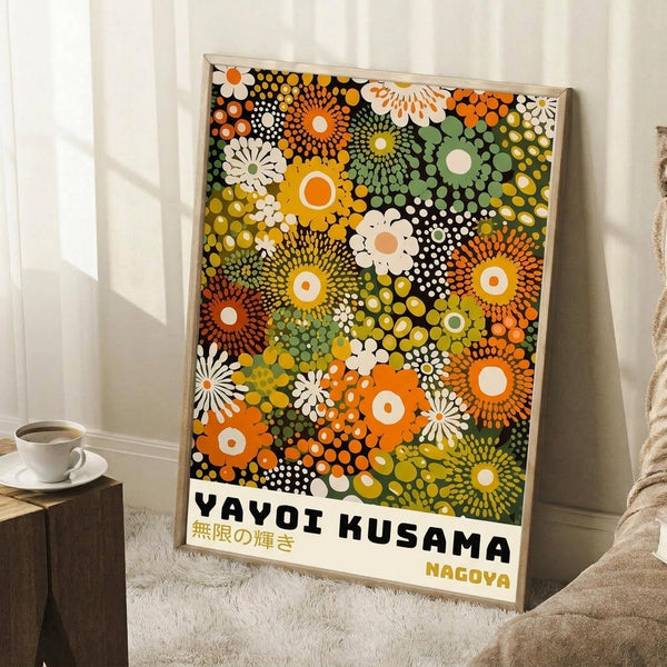 May include: Framed poster with vibrant psychedelic pattern of overlapping circular motifs in orange, yellow, green, and white on black background, featuring "YAYOI KUSAMA" and Japanese text at bottom. Leaning against white wall next to coffee cup on wooden block and beige throw blanket on white textured rug.