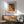 May include: Framed art print with colorful dotted pattern in orange, yellow, green and blue tones displayed above mid-century modern wooden sideboard with table lamp. Sage green upholstered armchair with wooden frame sits nearby in sunlit room with white walls and warm wood flooring.