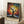 May include: Framed poster artwork featuring a black silhouette tree with sprawling branches against a vibrant background of colorful dots in blues, oranges, yellows, and reds resembling a starry night sky. Text reads "YAYOI KUSAMA" with Japanese characters below. The wooden-framed print leans against a white wall next to a wooden side table with white coffee cup and saucer, positioned on a light textured rug beside a beige blanket.